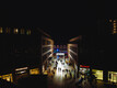 Eine Aufnahme der Kunstinstallation bei Nacht. Aus einer seitlichen Perspektive von oben leuchtet der Cube in der beschäftigten Fußgängerzone.